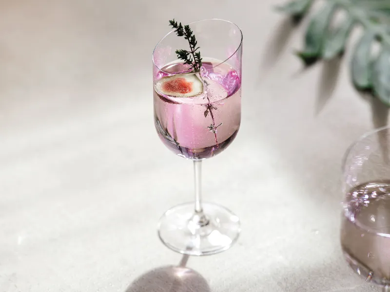 A pink cocktail, garnished with rosemary and fruit, is elegantly presented in a Villeroy & Boch NewMoon wine glass on a white background.