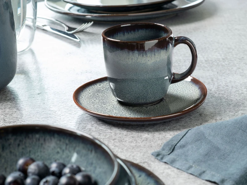 Lave Tasse auf einer passenden Untertasse, umgeben von ähnlichem Geschirr und einer Schale mit Heidelbeeren.