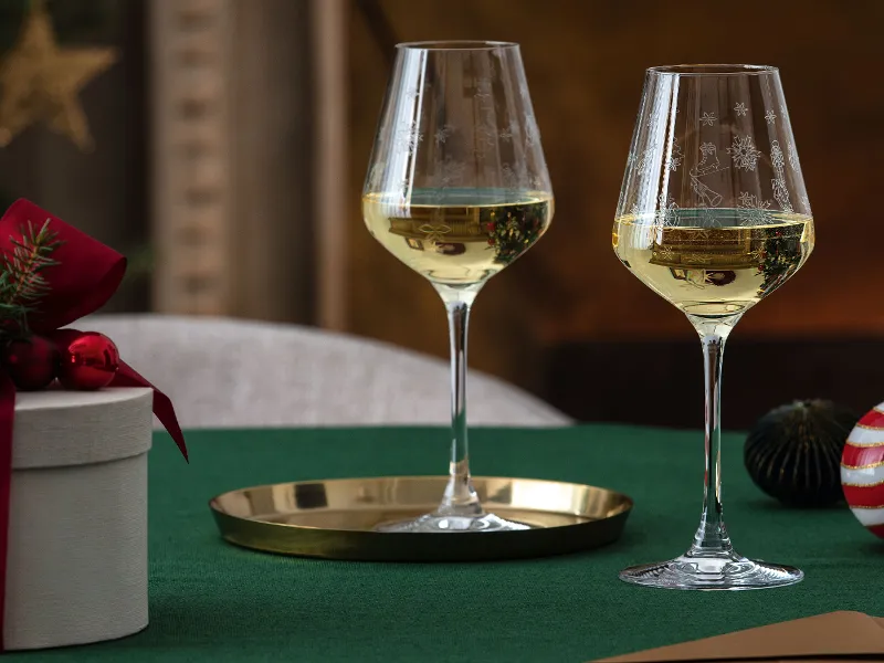 Two glasses of white wine on a green tablecloth are accompanied by a round white Toy’s Delight gift box from Villeroy & Boch with a red bow and festive decorations.