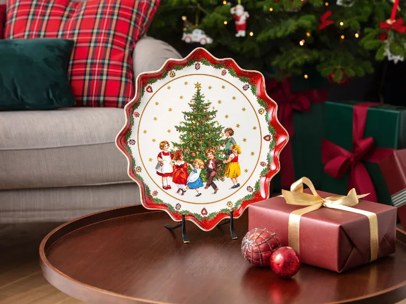 Une assiette festive avec des enfants autour d’un sapin de Noël est posée sur une table ronde en bois à côté d’un cadeau emballé et de boules décoratives. En arrière-plan, se trouvent une assiette Toy’s Fantasy de Villeroy & Boch et des coussins à carreaux.