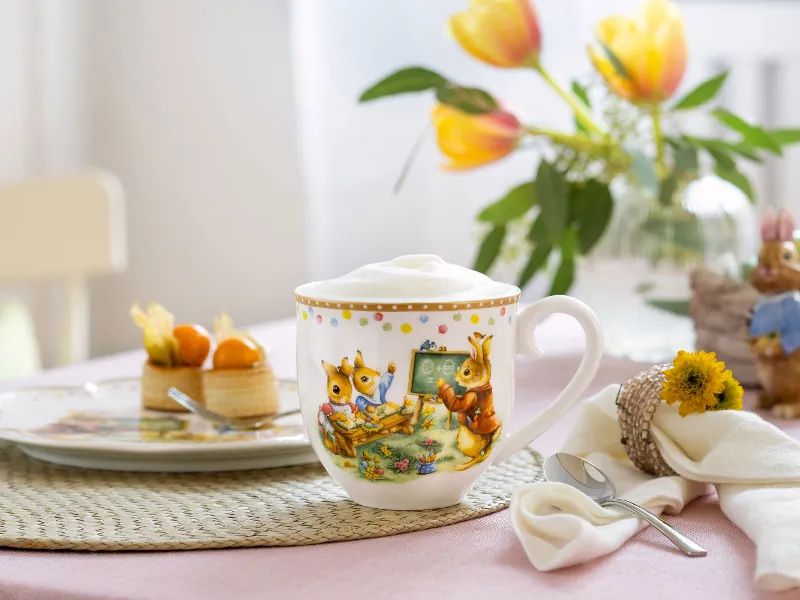 De decoratieve mok met schuimkroon uit de Villeroy & Boch Annual Easter Edition, gedecoreerd met een konijnenmotief, staat op een tafel naast een dessertbord, een gebloemd tafelkleed en een konijnenfiguurtje. Op de achtergrond bloeien tulpen.	