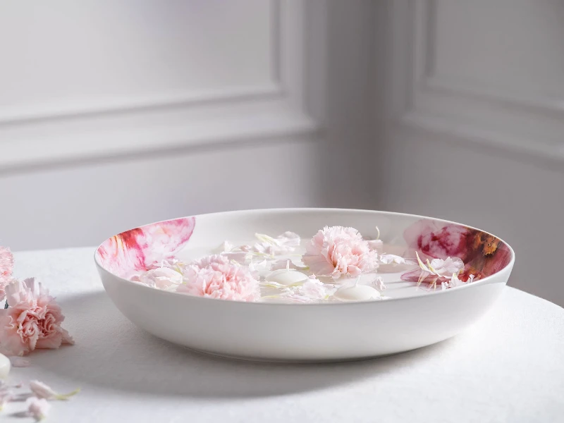 A Rose Garden serving bowl filled with water and floating pink flowers, surrounded by additional petals on a white surface.