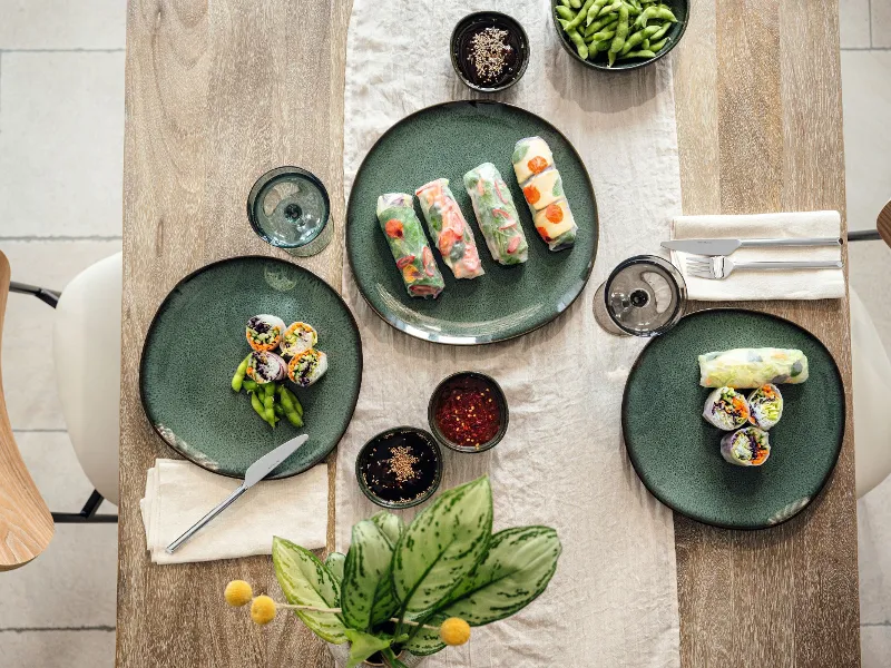 Top view of a wooden table with green crockery, on which sushi rolls, spring rolls, edamame and sauces are laid out.