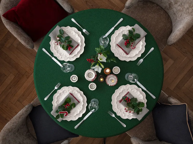 Une table ronde dressée en vert pour quatre personnes, décorée avec la vaisselle Toy’s Delight Royal Classic de Villeroy & Boch, des branches de houx, des bougies et des serviettes de fête.