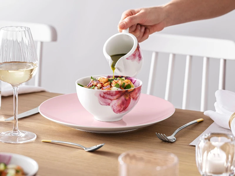 Une personne verse de la vinaigrette sur une salade dans un saladier Rose Garden, posé sur une table de salle à manger élégamment dressée avec de la vaisselle Rose Garden.