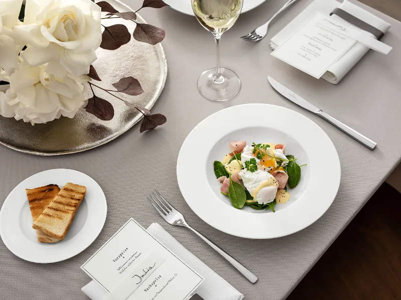 Composition de table élégante composée d'une assiette royale de Villeroy & Boch avec salade, complétée par un verre de vin blanc, une carte de menu et une garniture de pain grillé. Des roses blanches forment un point central élégant.	