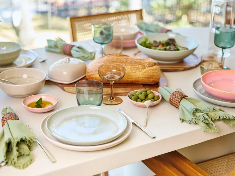 Tavolo apparecchiato in modo estivo con stoviglie della collezione Perlemor nei colori verde, beige e rosa, accompagnato da bicchieri e decorazioni nei toni del verde e del marrone.