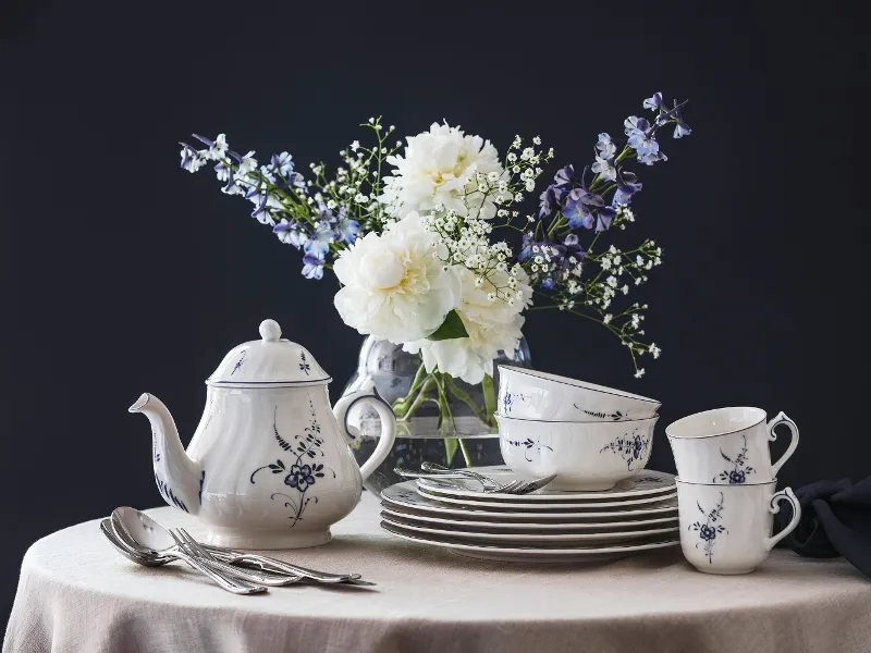 Een Vieux Luxembourg porseleinen servies met een blauw en wit bloemmotief, bestaande uit een theepot, kopjes en borden op een ronde tafel, met een vaas met witte en paarse bloemen op de achtergrond.