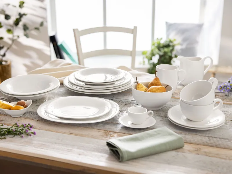 A New Cottage table setting by Villeroy & Boch featuring white tableware, cups, saucers, a bowl filled with pears, a jug, and a folded green napkin. A chair and plants can be seen in the background.