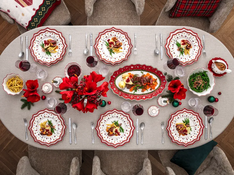 A festive dining table for six people with decorated plates, red flowers and bowls of food arranged in the centre.