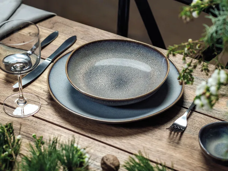 Une table est élégamment dressée avec la vaisselle beige « Lave » de Villeroy & Boch, accompagnée d'un verre à vin et de couverts posés sur une surface en bois, tandis que des plantes vertes et des fleurs blanches apportent une touche de décor.	