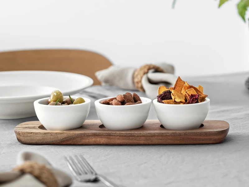 Three Artesano dip bowls with various snacks on a wooden serving tray, set on a table with elegant place settings and a stem with leaves.