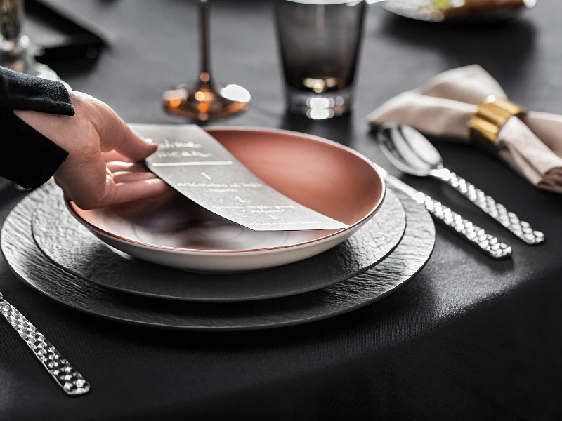 One person places a menu card on an elegant table with a black table cloth, flatware and glasses together with copper-colored and black Manufacture dinnerware.