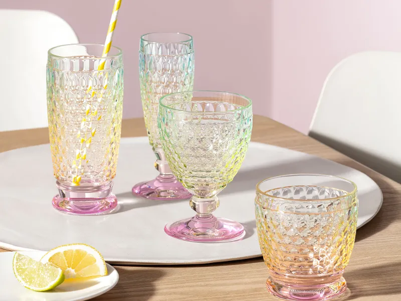 Four textured glasses from Villeroy & Boch Boston Pearl, three of them empty and one with a yellow-striped straw, stand on a white tray on a wooden table. Next to them is a small plate with lemon slices.