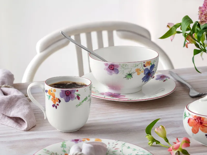 Die Mariefleur-Kollektion von Villeroy & Boch umfasst einen Becher, eine Schüssel und eine Untertasse mit Blumenmuster auf einem hellen Holztisch, begleitet von einer Serviette, einem Löffel und einem kleinen Blumenarrangement.