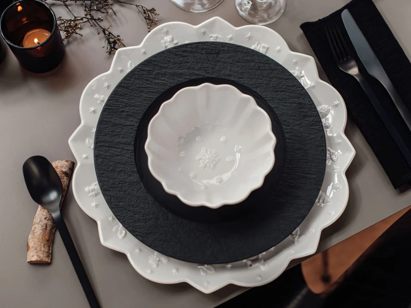 A table setting in black and white. It includes Villeroy & Boch Toy’s Delight Royal Classic plates with scalloped edges and round plates, a white bowl, black flatware, a napkin, and three lighted black candles.