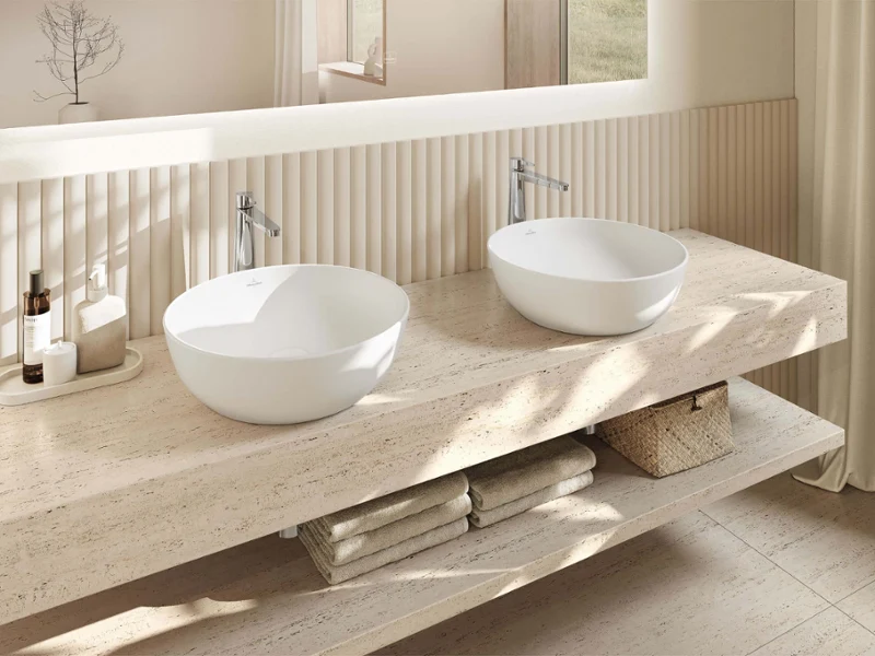 A modern bathroom with two round Artis washbasins on a marble countertop, a mirror, toiletries, folded towels and a storage basket underneath.