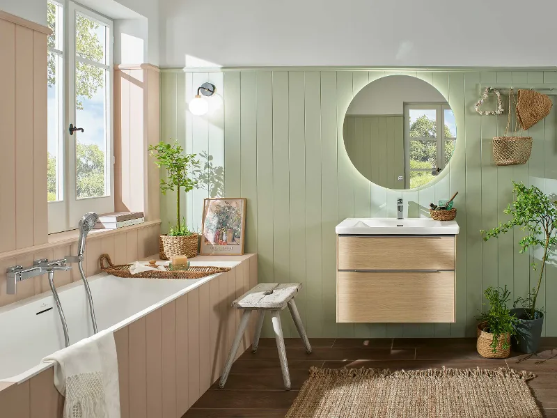 Badkamer met een Subway 3.0 bad van Villeroy & Boch, ronde spiegel en houten wastafel. Planten en rieten decoraties voegen een natuurlijk tintje toe. Houten lambrisering in pastelkleuren onderstreept de rustieke esthetiek. Licht stroomt naar binnen door het raam.