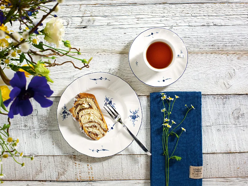 Una tazza da tè Vieux Luxembourg con pasticcini su un piatto, un libro e una composizione floreale su un tavolo di legno rustico.