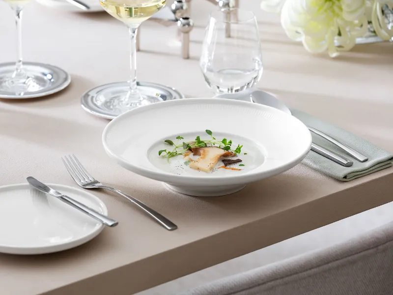 Une assiette creuse Afina de Villeroy & Boch contenant une soupe de cèpes à la crème, à côté d’un verre à vin blanc ainsi que des couverts de table Piemont.