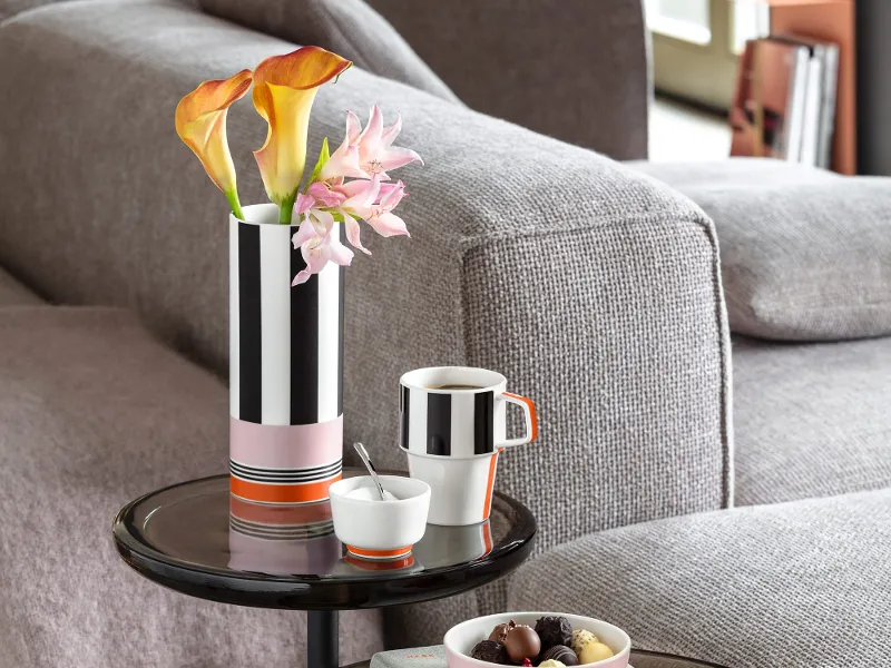 Two round side tables with a Memphis flower vase, a Memphis coffee cup, a sugar bowl, a book and a bowl of chocolates in a modern living room.