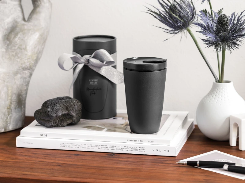 A stylish workspace with decorative objects, including two black Manufacture To Go coffee mugs, a white vase with blue flowers, stacked books and a stone sculpture on a wooden surface.