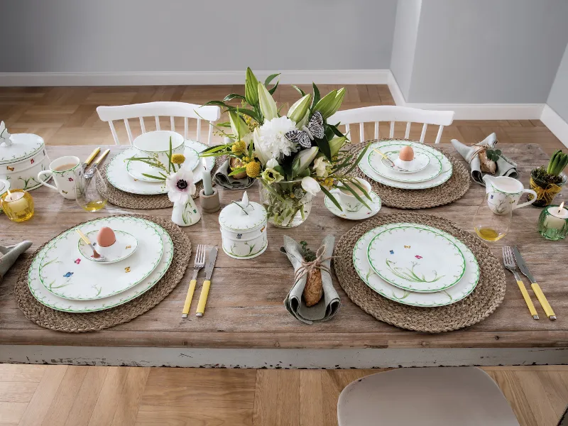 Une présentation de table « Colourful Spring » de Villeroy & Boch pour quatre personnes se composant de porcelaine à motifs floraux, de couverts de table à manches jaunes, de sets de table tissés et d’une composition florale. Chaque couvert dressé est accompagné d’un œuf dans une tasse décorative.