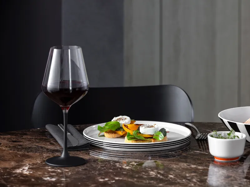 A dining room with a glass of red wine, Memphis plates with appetizers and two Memphis bowls on a marble table.
