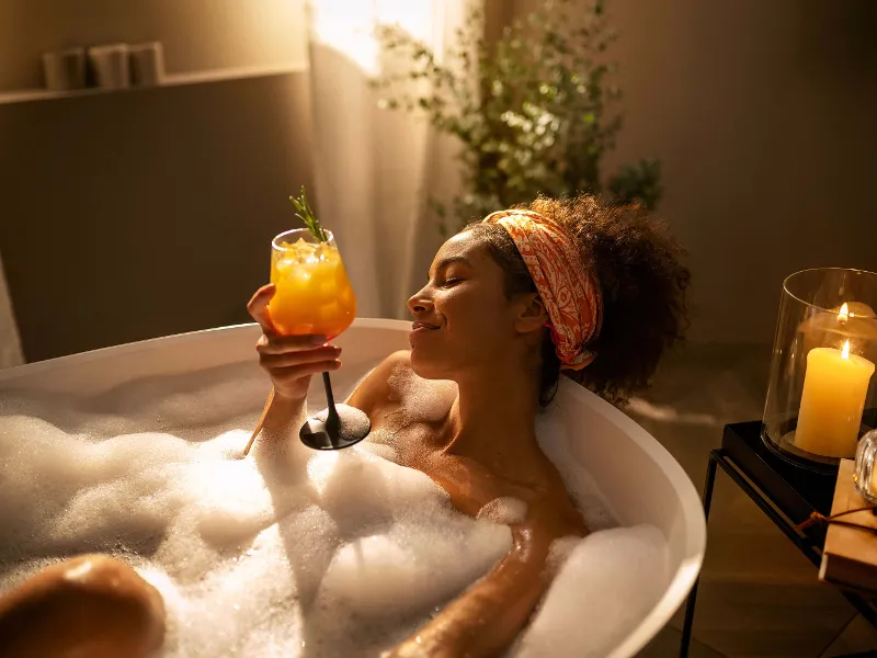 Une femme, une boisson à la main, est allongée dans une baignoire éclairée par des bougies.