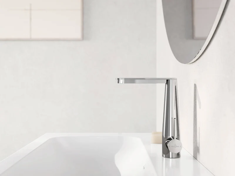 A modern chrome tap extends over a white washbasin, with part of a mirror and a beige wall in the background.