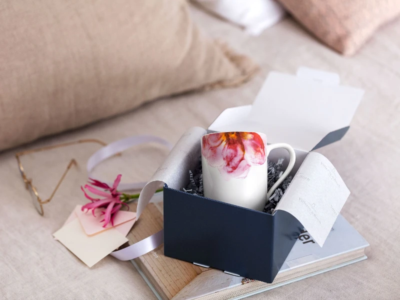 Eine offene Villeroy & Boch Geschenkbox mit einer Rose Garden Tasse, rosa Blumen und einer Notiz auf einem Holztablett, im Hintergrund weiche Kissen.