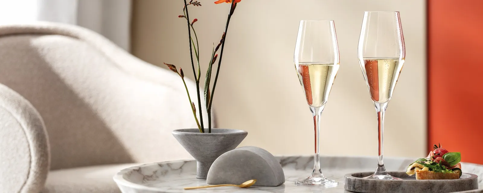 Une paire de verres La Divina de Villeroy & Boch est posée sur une table en marbre, accompagnée de fleurs orange dans un vase et d’un canapé, créant une ambiance élégante dans un salon plein de style.