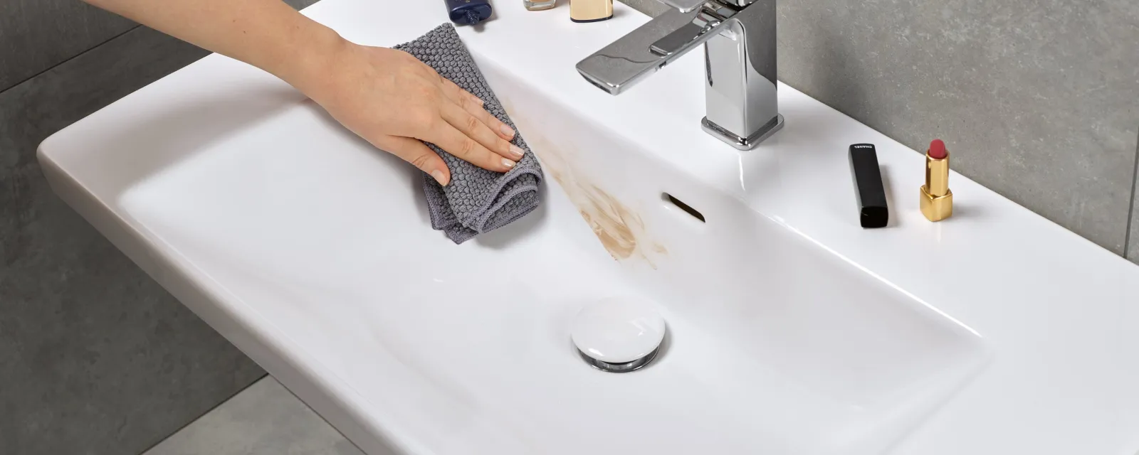 Czyszczenie zabrudzonej umywalki Ręka czyszcząca szarą ściereczką umywalkę w łazience i usuwająca plamę z produktu do makijażu. Na krawędzi umywalki znajdują się różne produkty do makijażu.
