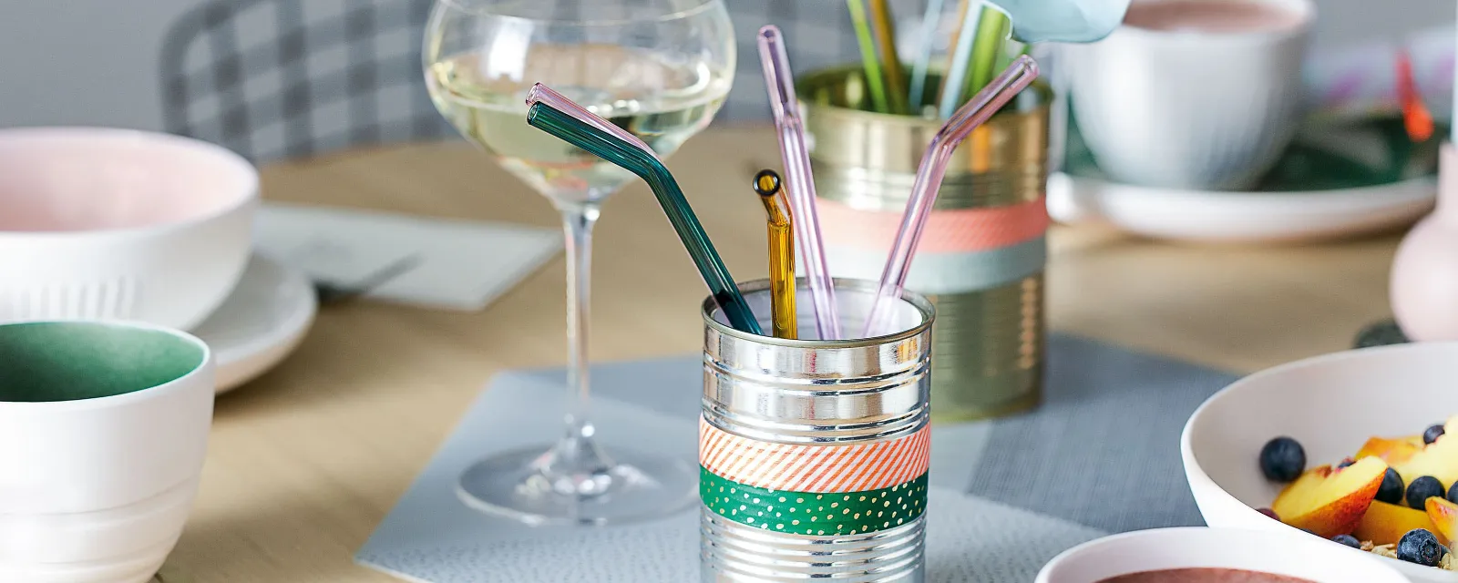 Elegant table setting with Artesano glass straws Elegantly set dining table with a glass of white wine, colorful straws in a decorative holder, and bowls of fresh fruit and a smoothie.