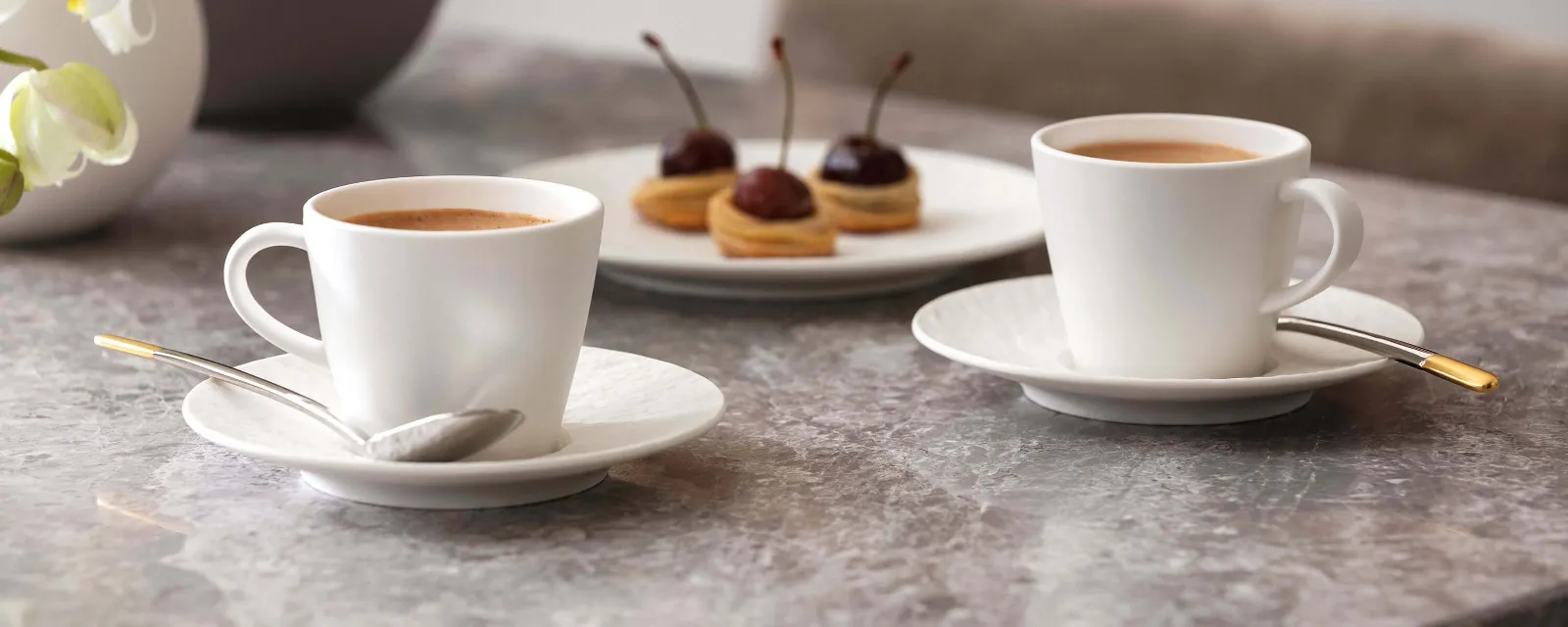 Two Manufacture espresso cups and a plate of pastries on a marble table with orchids in the background.