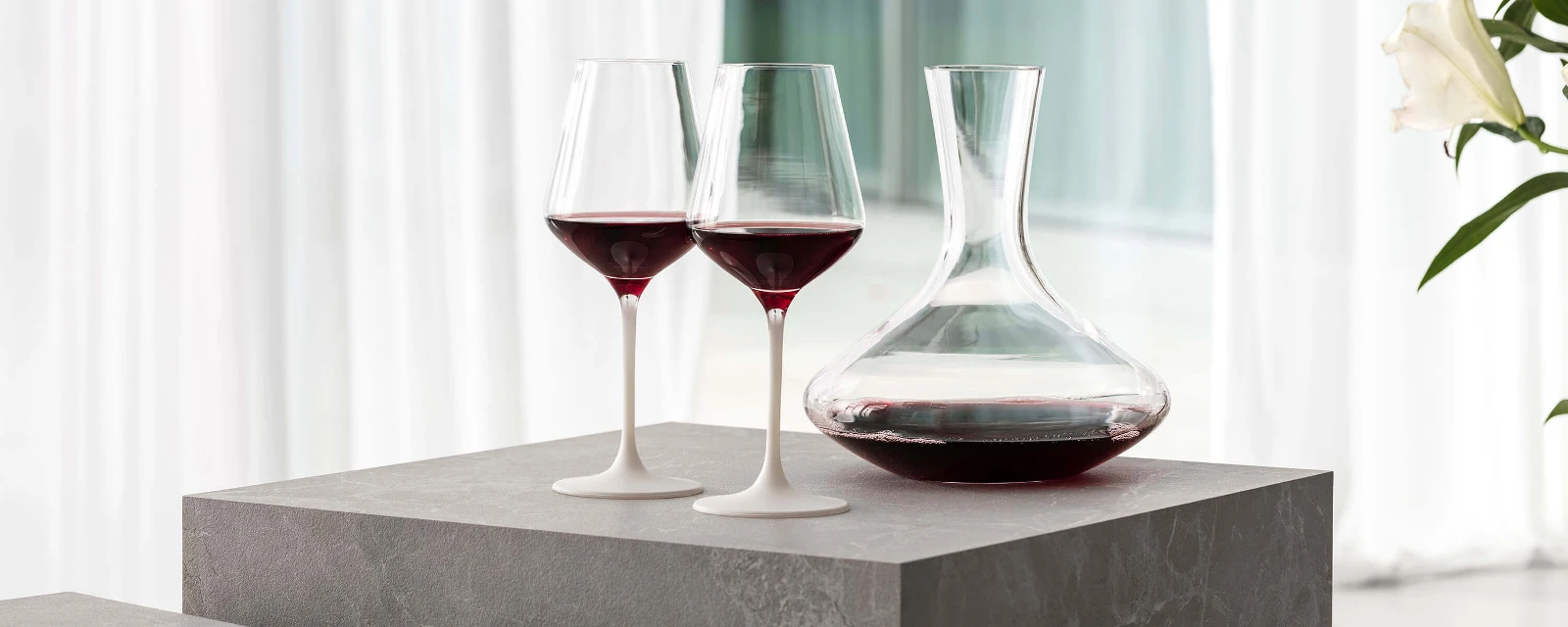 Two filled Manufacture red wine glasses on a table next to an elegant Purismo wine decanter.