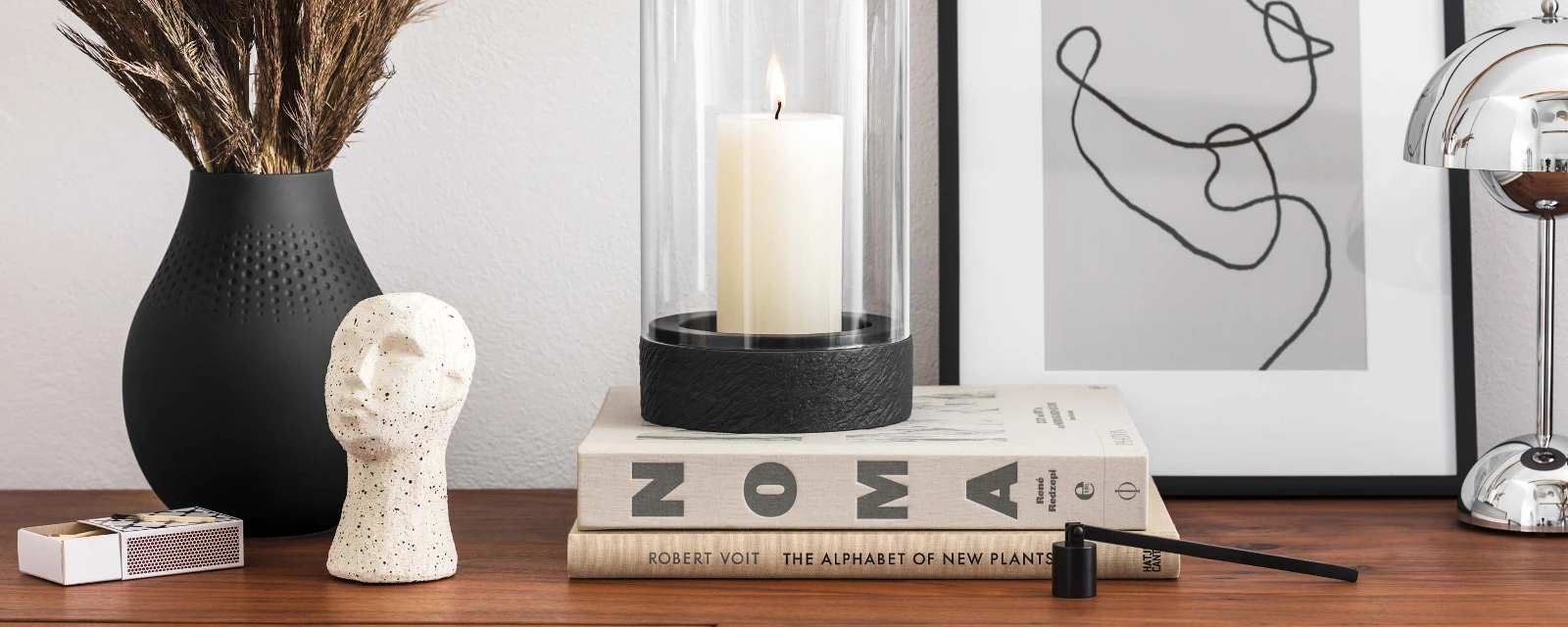 A modern highboard with decorative objects, including a vase with dried plants, a black Manufacture hurricane lamp, a sculpture, stacked books and a piece of framed abstract artwork.