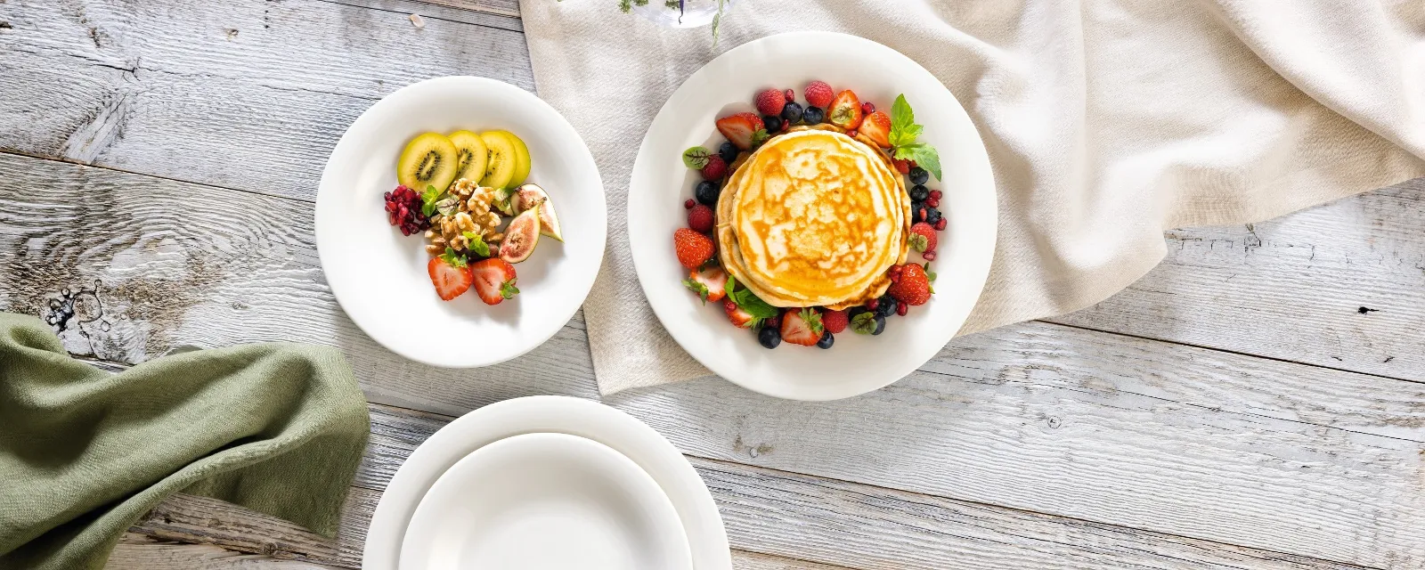 Une pile de crêpes aux baies est servie sur une assiette New Cottage de Villeroy & Boch, tandis que des fruits coupés sont servis sur une autre assiette. La table en bois est décorée avec des fleurs et une nappe drapée.