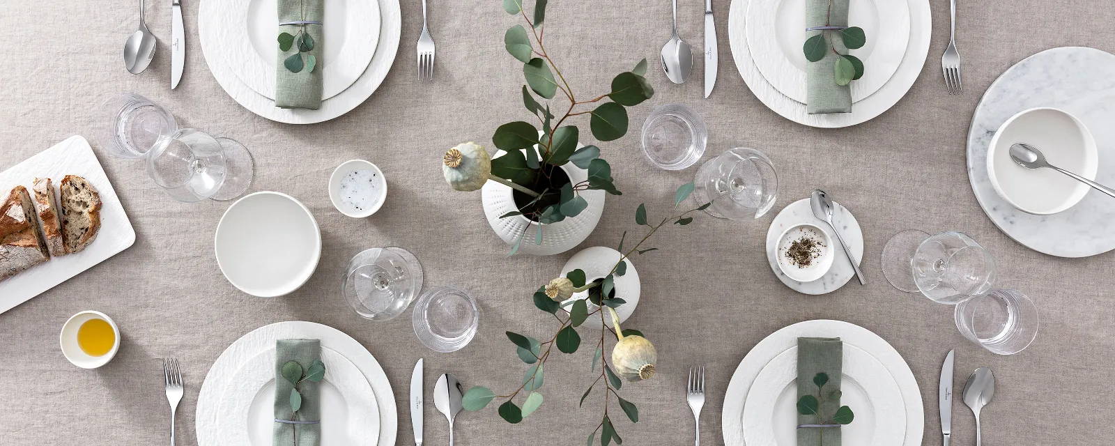 Une table dressée avec une nappe beige, des assiettes blanches Manufacture, des couverts de table, des verres à vin et une composition florale