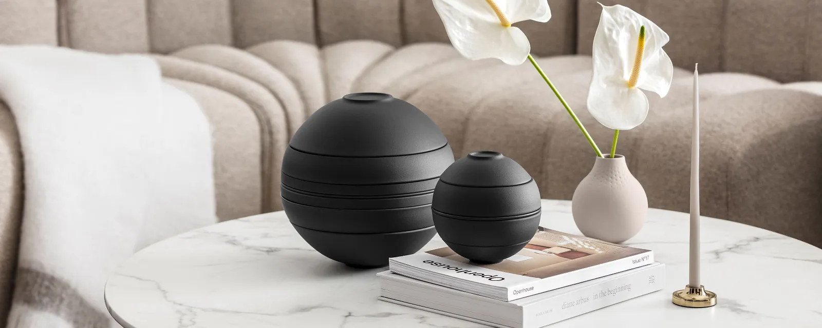 La Boule noire de Villeroy & Boch dans une ambiance de salon beige La Boule noire de Villeroy & Boch, un vase beige avec des fleurs blanches, un bougeoir et quelques livres dans une ambiance de salon beige.