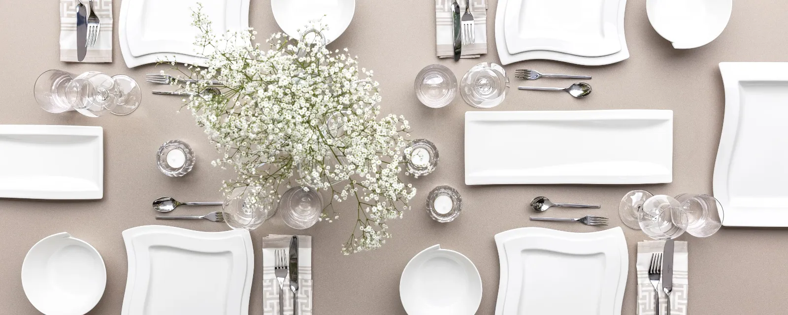 The Villeroy & Boch NewWave table set consisting of square white plates, bowls, cutlery, glasses and a centrepiece with white flowers on a beige tablecloth for four people.