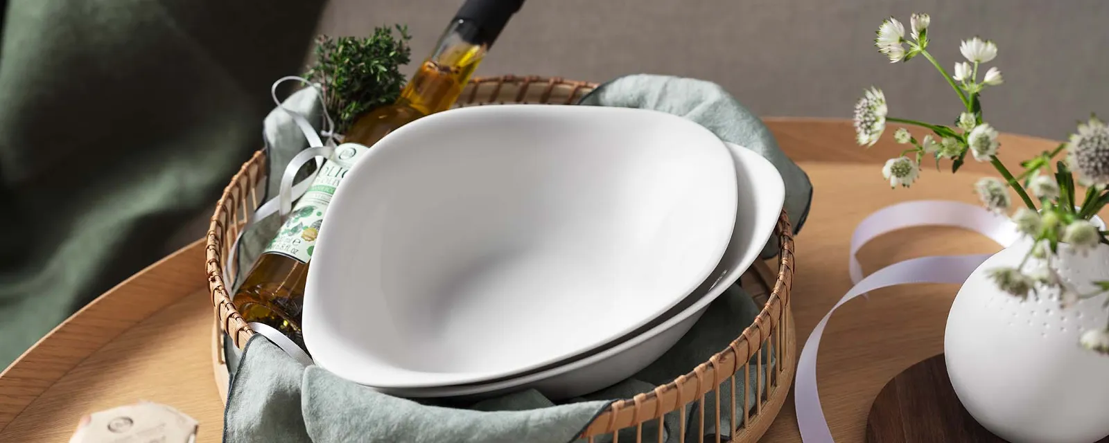 Des assiettes Vapiano dans une corbeille, avec une bouteille d’huile sur une table en bois, à côté d’un vase et de papier à lettres de Villeroy & Boch