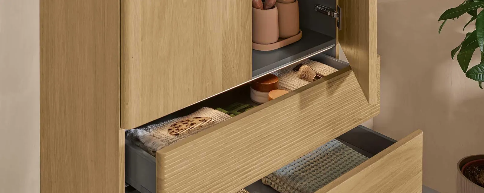 Une armoire de salle de bains Villeroy & Boch en bois clair avec deux portes et deux tiroirs ouverts adossés à un mur naturel.