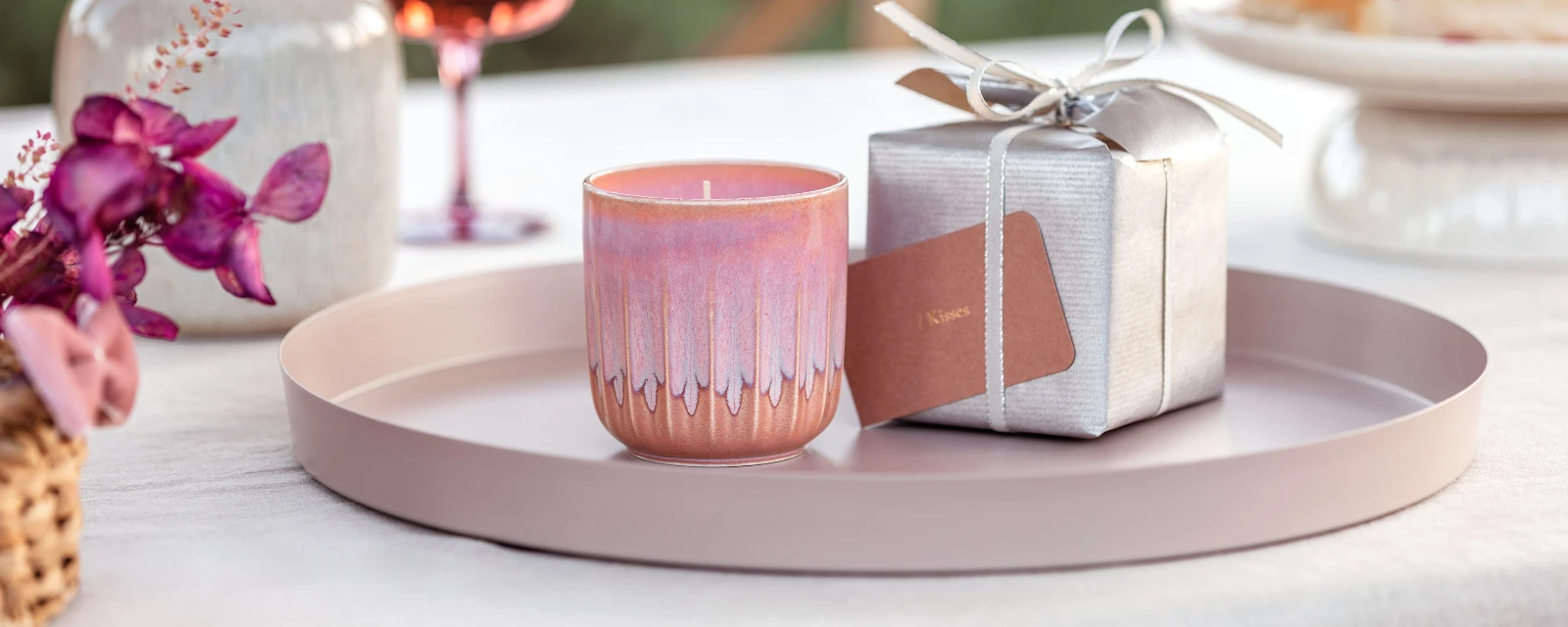 Scented candle from the like. by Villeroy & Boch collection as a gift on a round tray with a silver gift box in the background and pink orchids in the foreground.