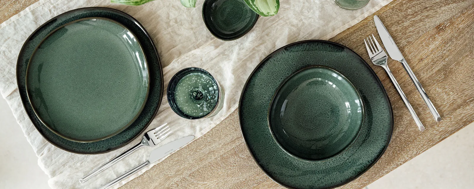 Stylishly set wooden table with "Lave Vert" ceramic tableware, silver "like cutlery" from like. by Villeroy & Boch, and matching Like glasses on a cream-coloured fabric napkin. A green plant with yellow accents complements the elegant arrangement.