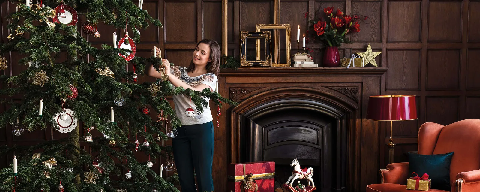 Weihnachtsbaum geschmückt mit Toy's Delight Tellern und Ornamenten Eine Frau schmückt einen Weihnachtsbaum in einem gemütlichen Wohnzimmer. Unter dem Baum liegen verschiedene verpackte Geschenke. Der Raum ist mit Holzvertäfelungen, einem Kamin und einem roten Sessel geschmückt.