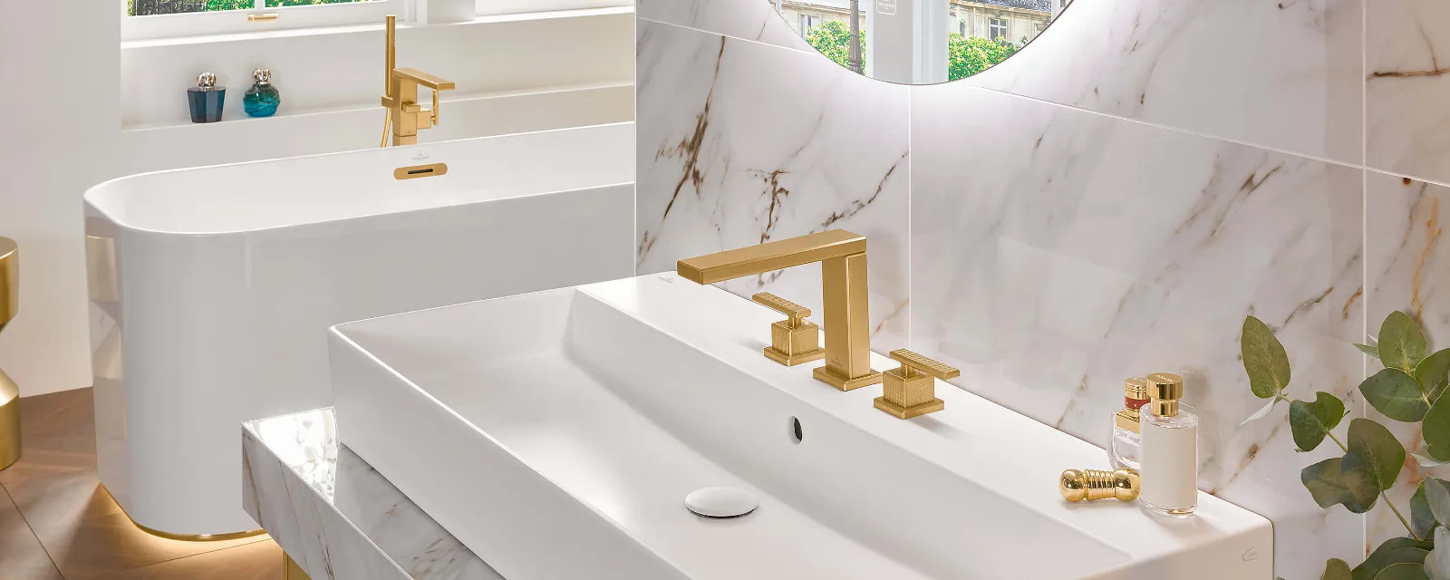 Une salle de bains moderne avec des surfaces en marbre, un grand lavabo rectangulaire, des robinetteries dorées, un miroir rond, une baignoire en îlot et une plante verte en guise de décoration. La lumière naturelle passe à travers les fenêtres.