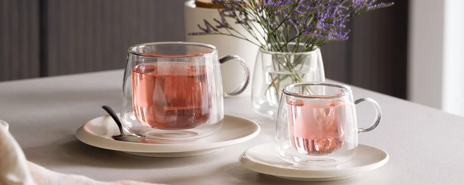 Des tasses en verre Artesano de Villeroy & Boch remplies de thé rose, placées sur des soucoupes à côté d'une cuillère sur une table décorée d'un petit vase de fleurs violettes.