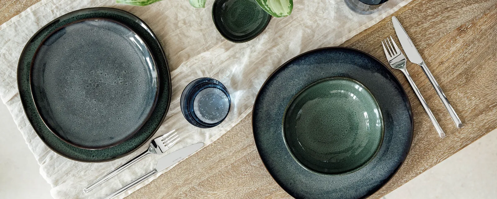 Lave place settings with green plates, cutlery and blue glasses decorate a wooden table Two Villeroy & Boch Lave place settings with green plates, cutlery and blue glasses adorn a wooden table with a white table runner. In the centre is a green-leaved plant in a vase.