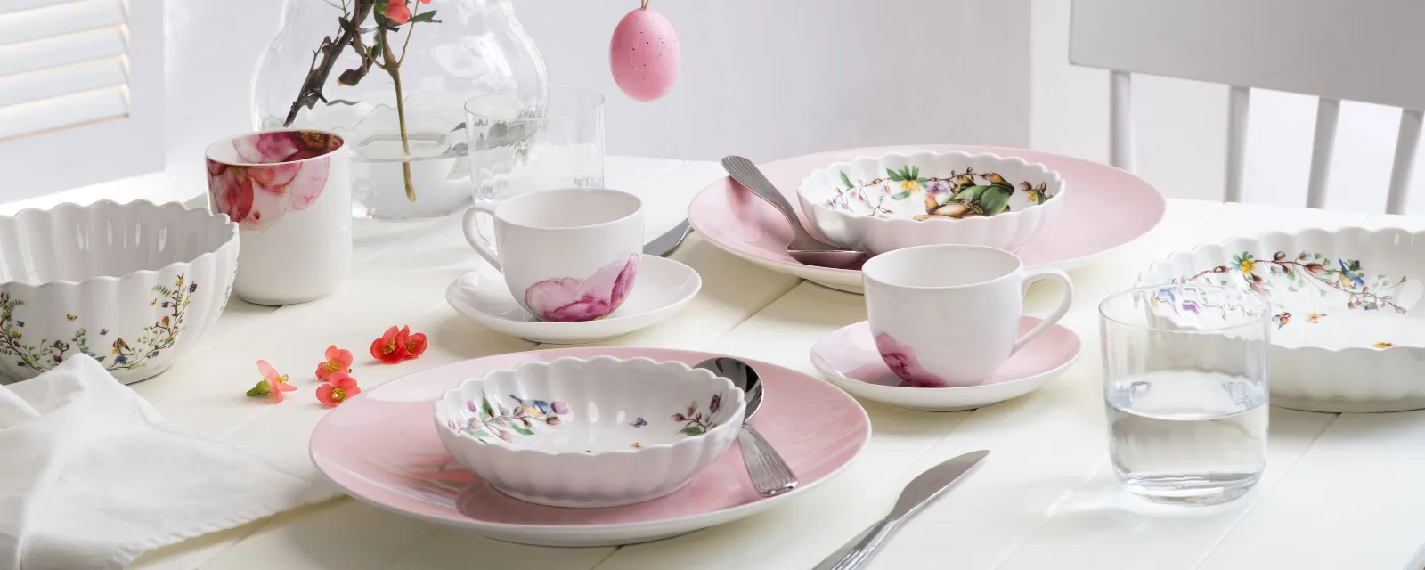 A bright, spring-like dining table with pastel-coloured tableware from the Rose Garden and Spring Fantasy collections, decorated with floral patterns and Easter eggs hanging from a branch as a centrepiece.